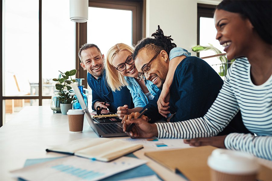 About - Group of Peers Smiling and Laughing While Sitting at a Wooden Table with Office Supplies and a Computer While Light Shines Through a Window